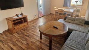 A living room with a wooden floor, a glass coffee table, a sofa, a TV mounted on the wall, and a dining area with a table and chairs. - Hotel Bischoff (Winnweiler)