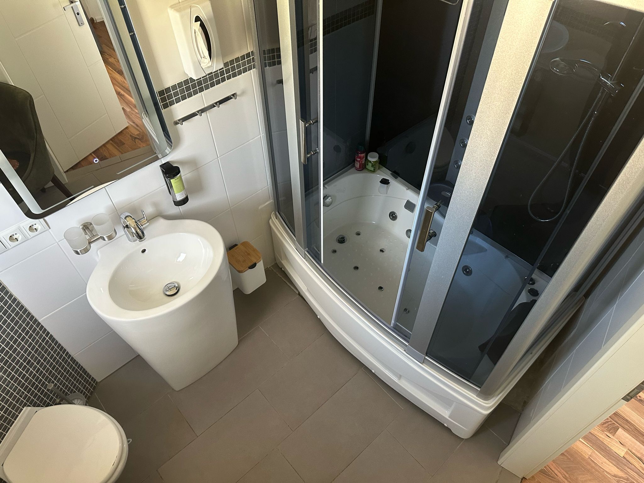 Un baño moderno con un lavamanos blanco, un espejo, un cabezal de ducha manual y una ducha con mampara de vidrio.