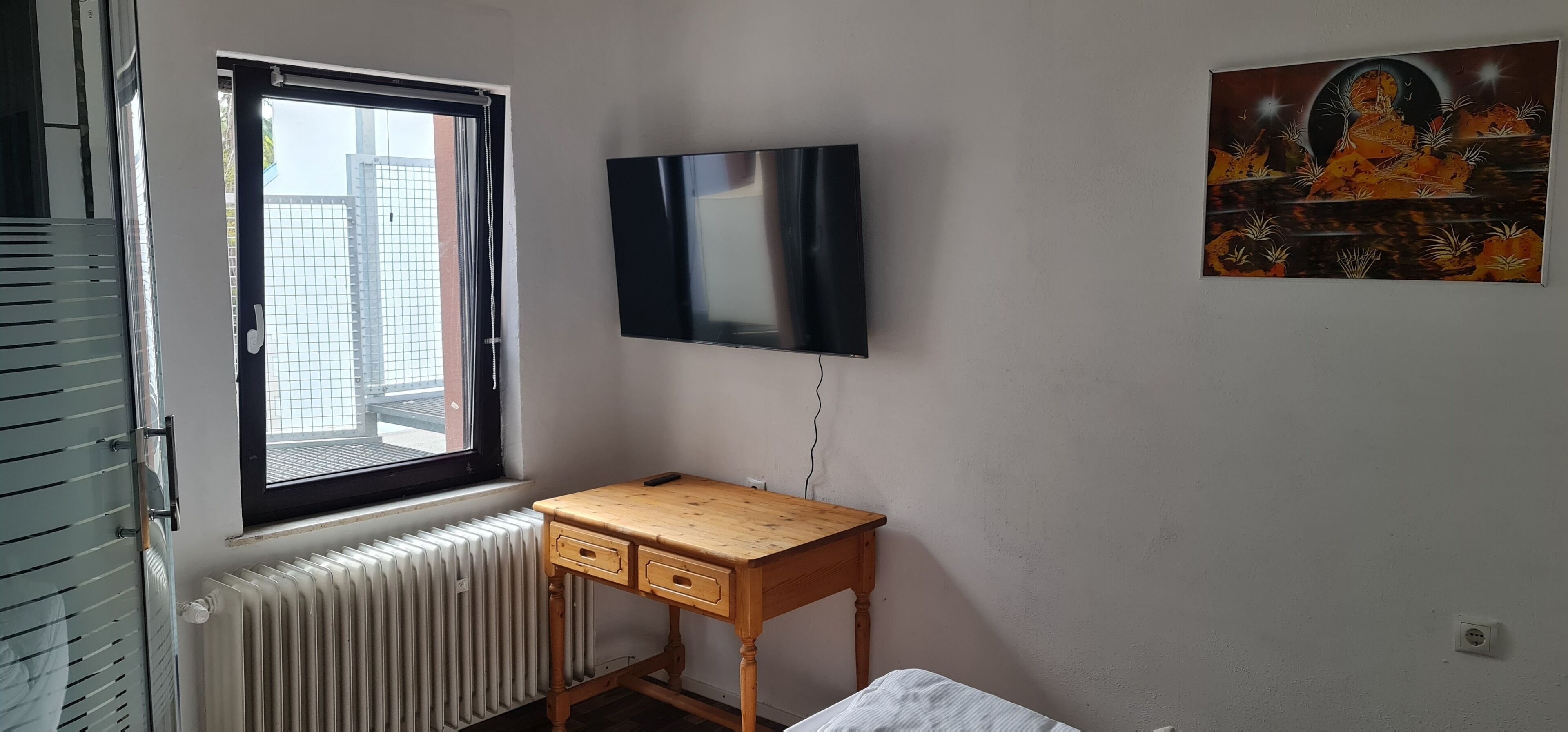 Ein Zimmer mit Fenster, Heizkörper, Schreibtisch aus Holz, Fernseher und einem Bild an der Wand.