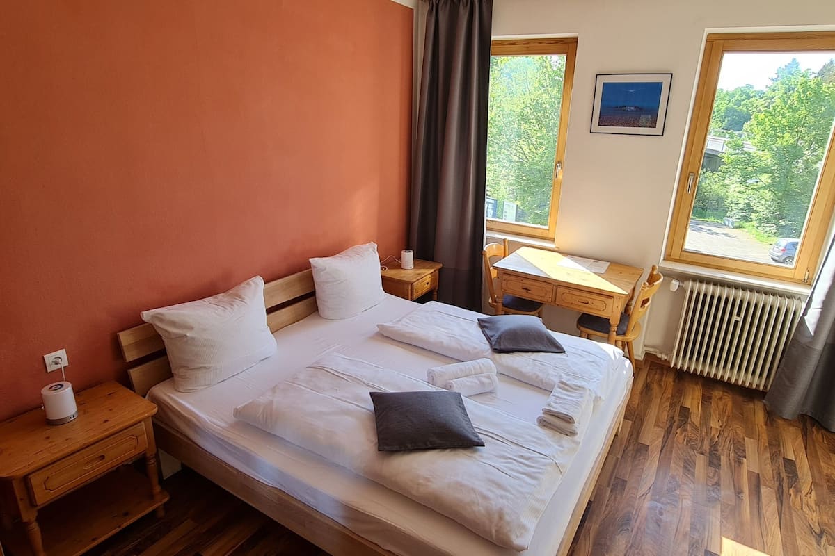 A bedroom with a wooden bed, two bedside tables, a window with a view of greenery, and a radiator.