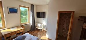 Une chambre avec un bureau en bois, un lit, une fenêtre avec des rideaux, un radiateur, une télévision et une salle de bain avec du papier peint à motifs.