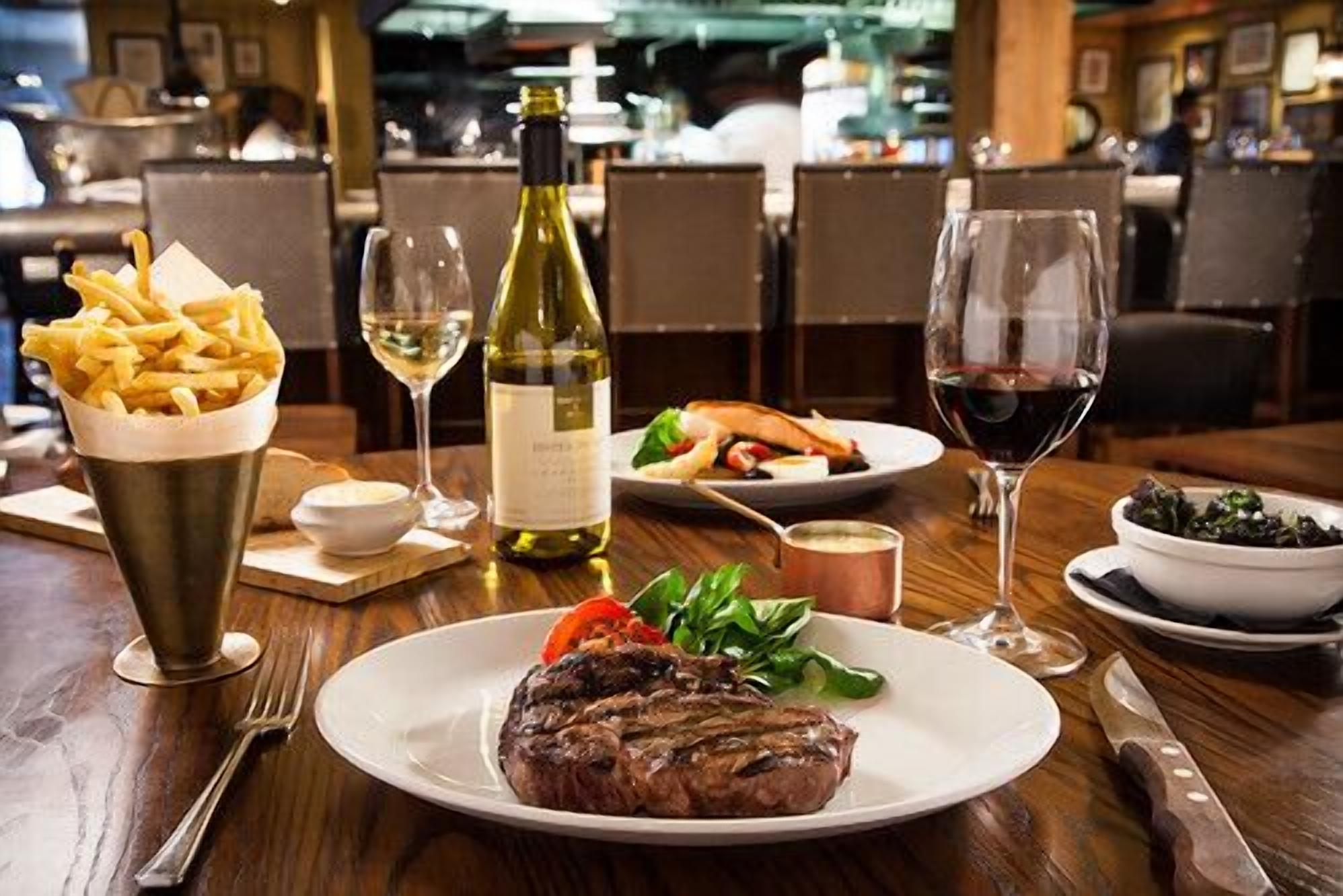 Een eettafel met een steak, frietjes, salade, wijn en een fles wijn.