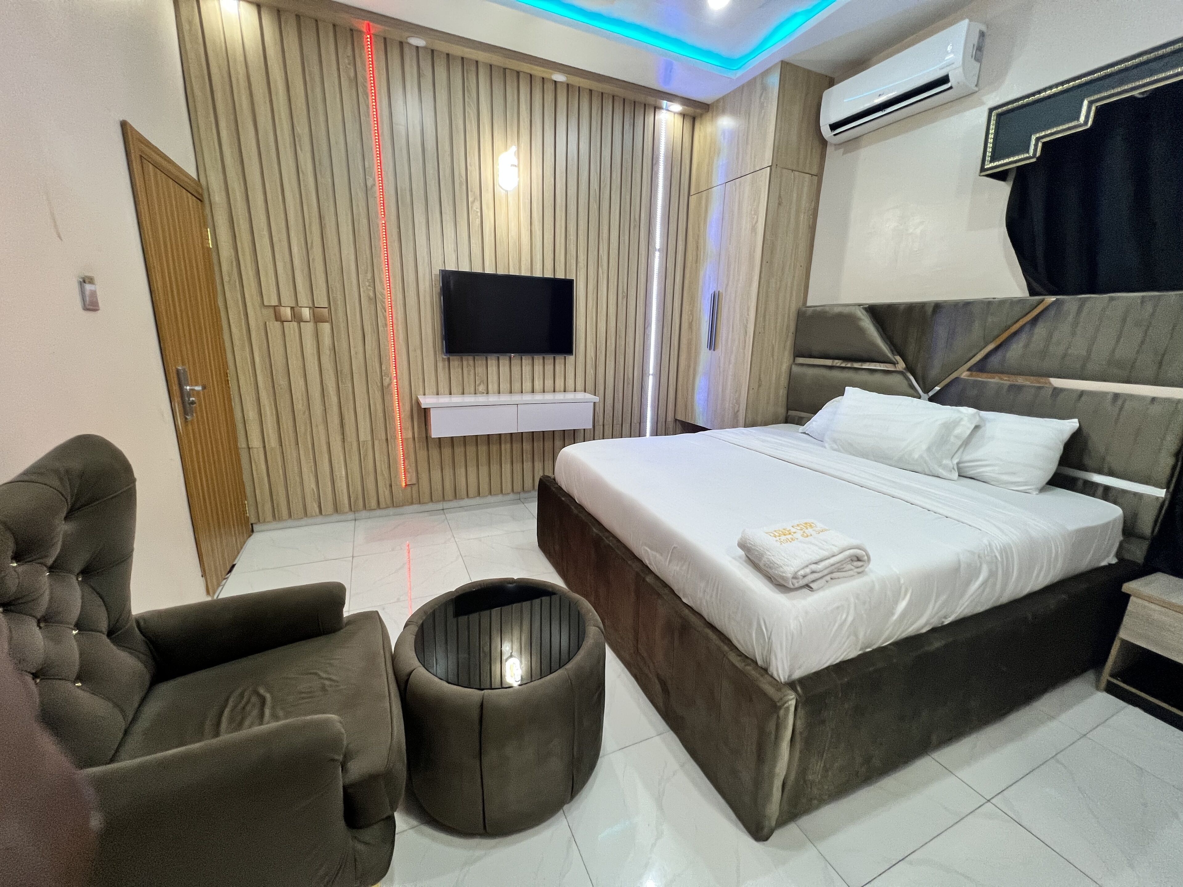 A modern bedroom with a large bed, a flat-screen TV, a wooden panel wall, and a ceiling light fixture.