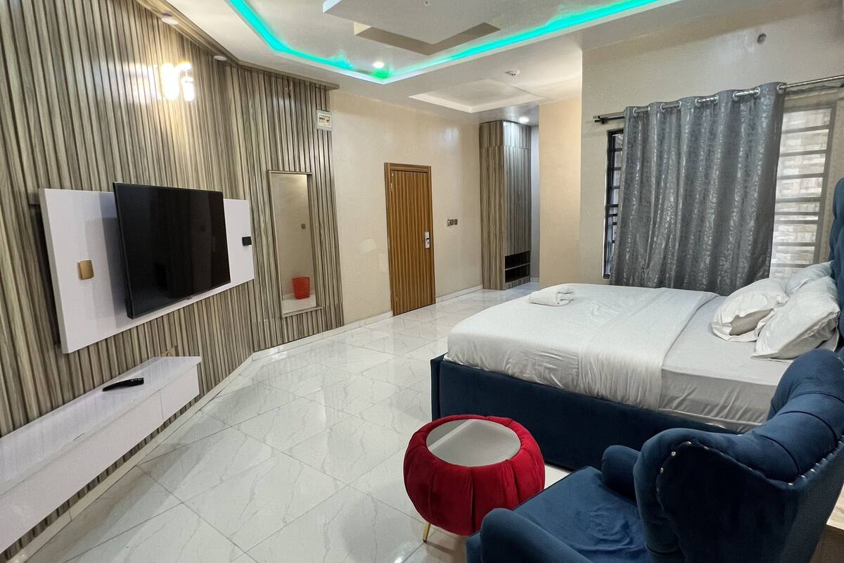 Ein modernes Schlafzimmer mit einem großen Bett, einem an der Wand montierten Fernseher und einem roten Hocker.