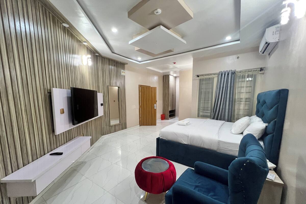 Ein modernes Hotelzimmer mit einem großen Bett, einem an der Wand montierten Fernseher, einem roten Pouf und einem blauen Sessel.