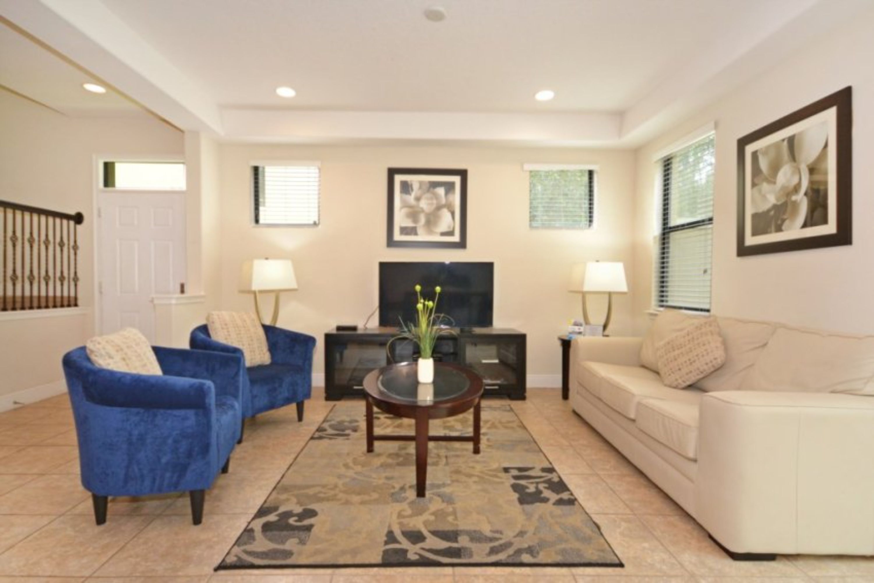 A living room with a beige sofa, a blue armchair, a coffee table with a vase, a TV, and two lamps.