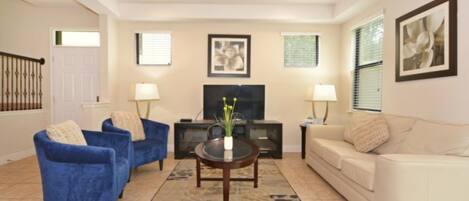 A living room with a beige sofa, a blue armchair, a coffee table with a vase, a TV, and two lamps.