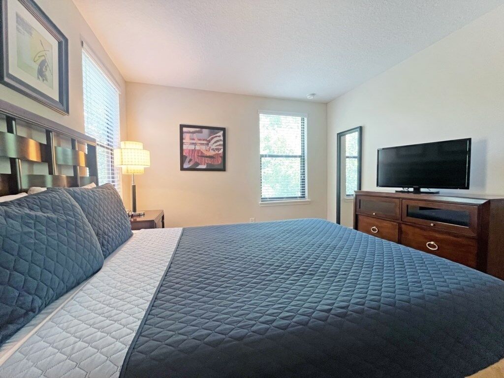 A hotel room with a large bed, a television, a dresser, and a window with blinds.