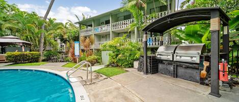 A pool area with a grill and lounge chairs, surrounded by lush greenery and a multi-story building with balconies.