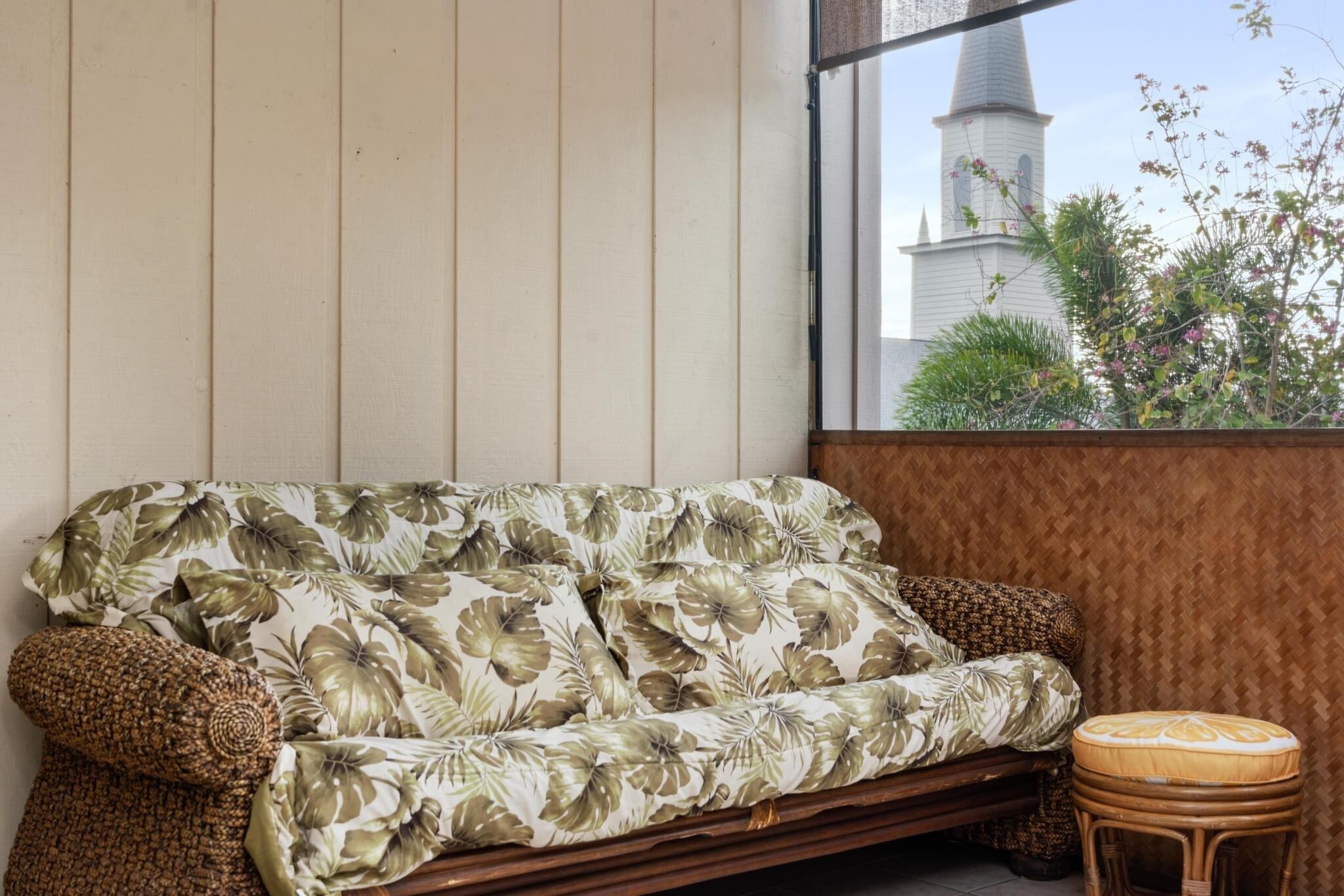 A room with a sofa and a small stool, a window with a view of a church steeple, and a wooden wall.