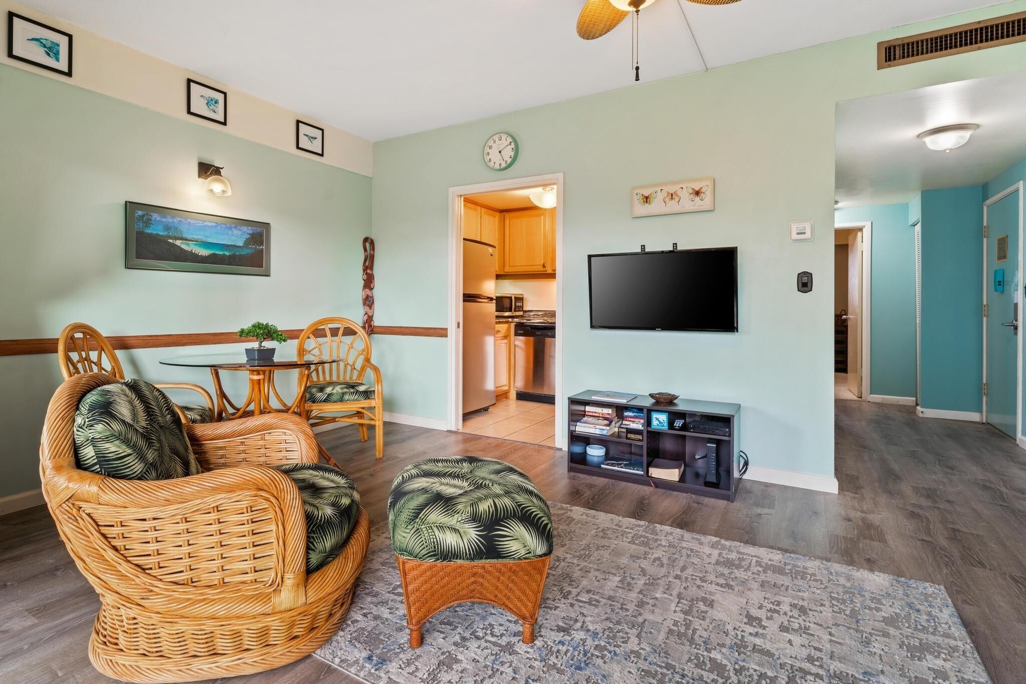 A living room with wicker furniture, a flat-screen TV, and a kitchen visible through an open doorway.