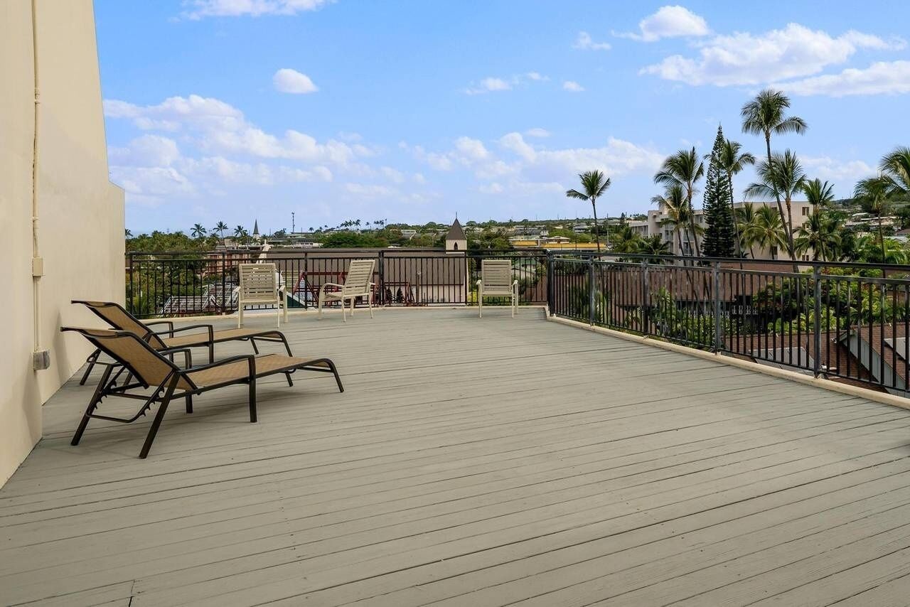 A wooden deck with lounge chairs, a metal railing, and a view of palm trees and buildings in the distance.