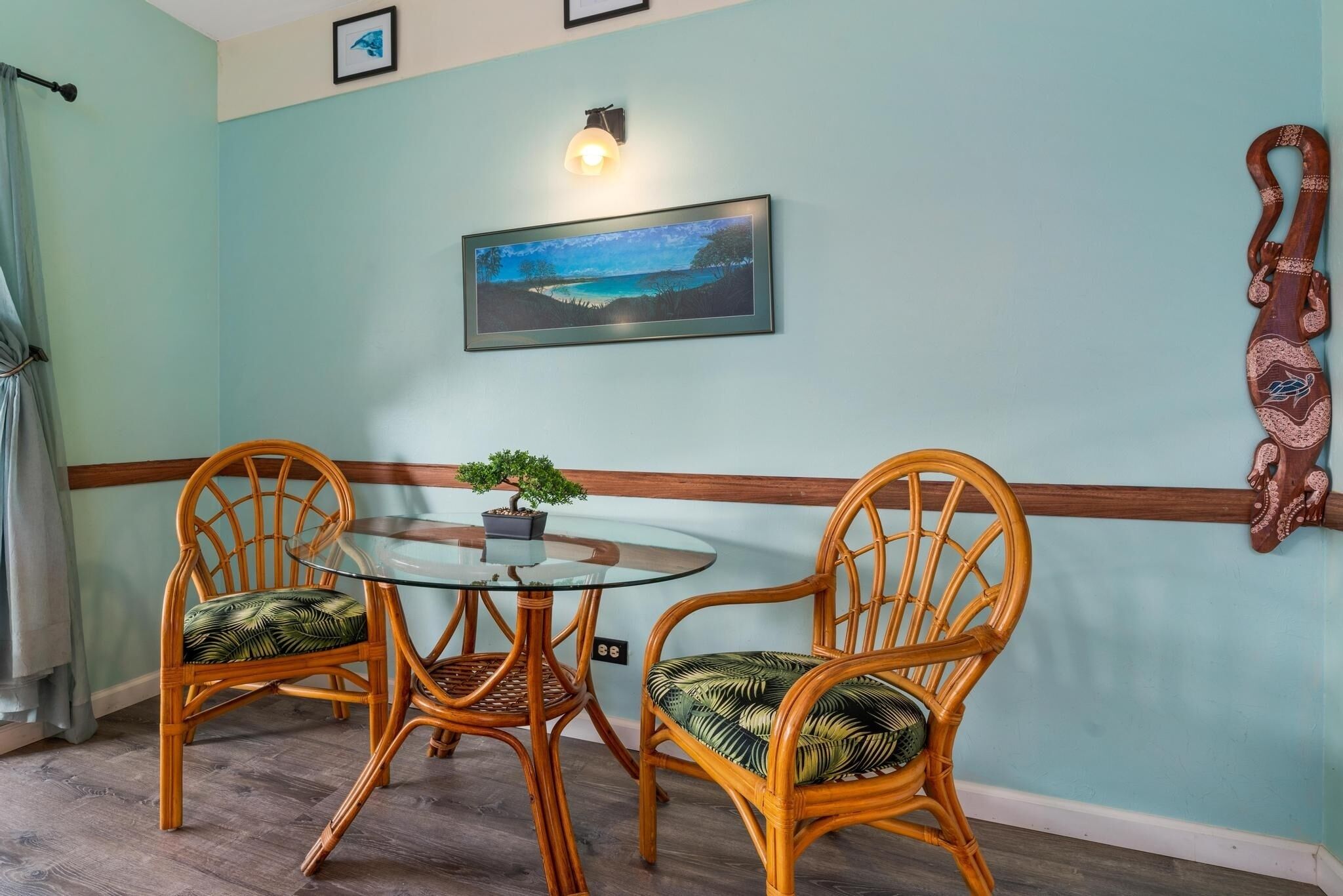 Un coin repas avec deux chaises en osier et une table en verre, un tableau encadré accroché au mur et une petite plante en pot posée sur la table.