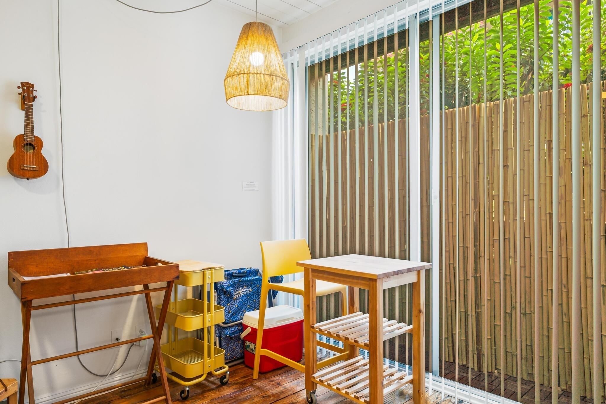 A room with a wooden table, a chair, a small guitar, a hanging light fixture, and a bamboo fence outside.