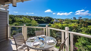 A balcony with a round table set for dining, surrounded by chairs, overlooking a lush garden and distant buildings.