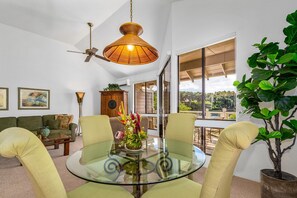 A modern living room with a glass table, green chairs, a ceiling fan, and a large window offering a view of greenery. - Grand Champions One Bedrooms by Coldwell Banker Island Vacations (Kihei)