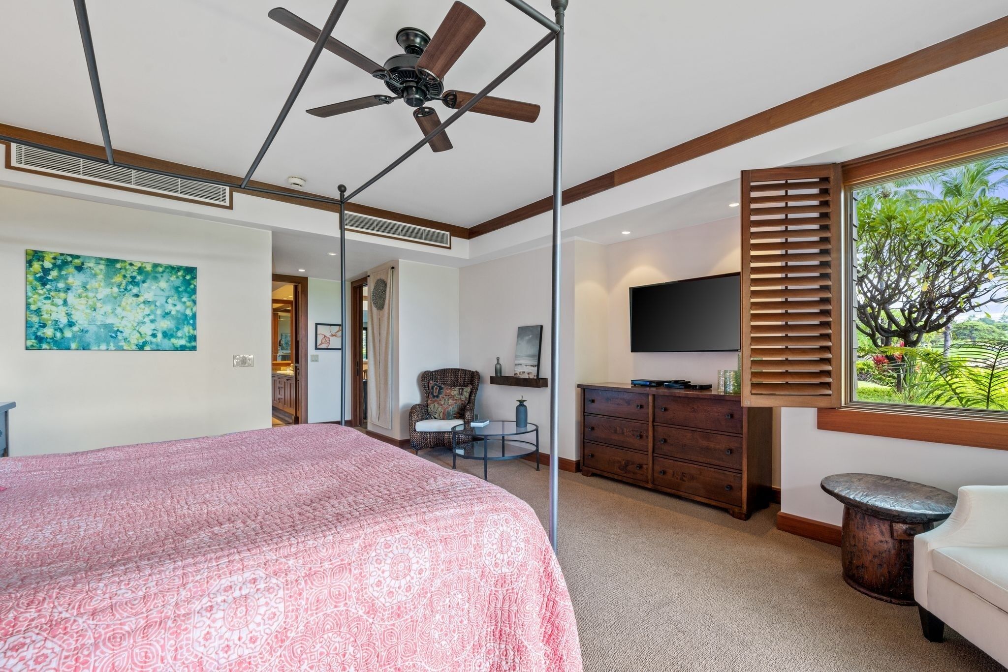 A bedroom with a large bed, a ceiling fan, a TV, a dresser, and a window with a view of greenery.