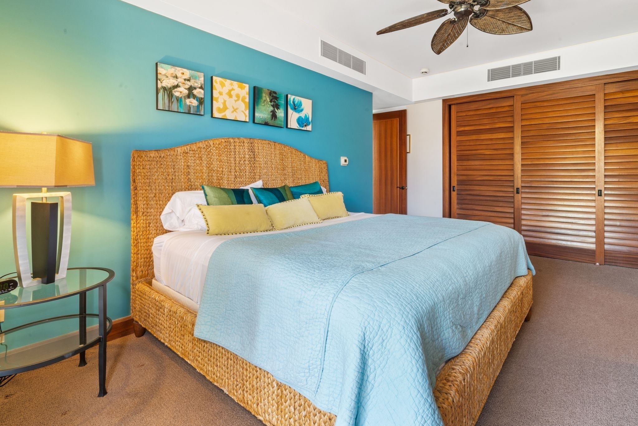Un dormitorio con cabecera de mimbre, cama con colcha azul, mesita de vidrio con lámpara y armario de madera.