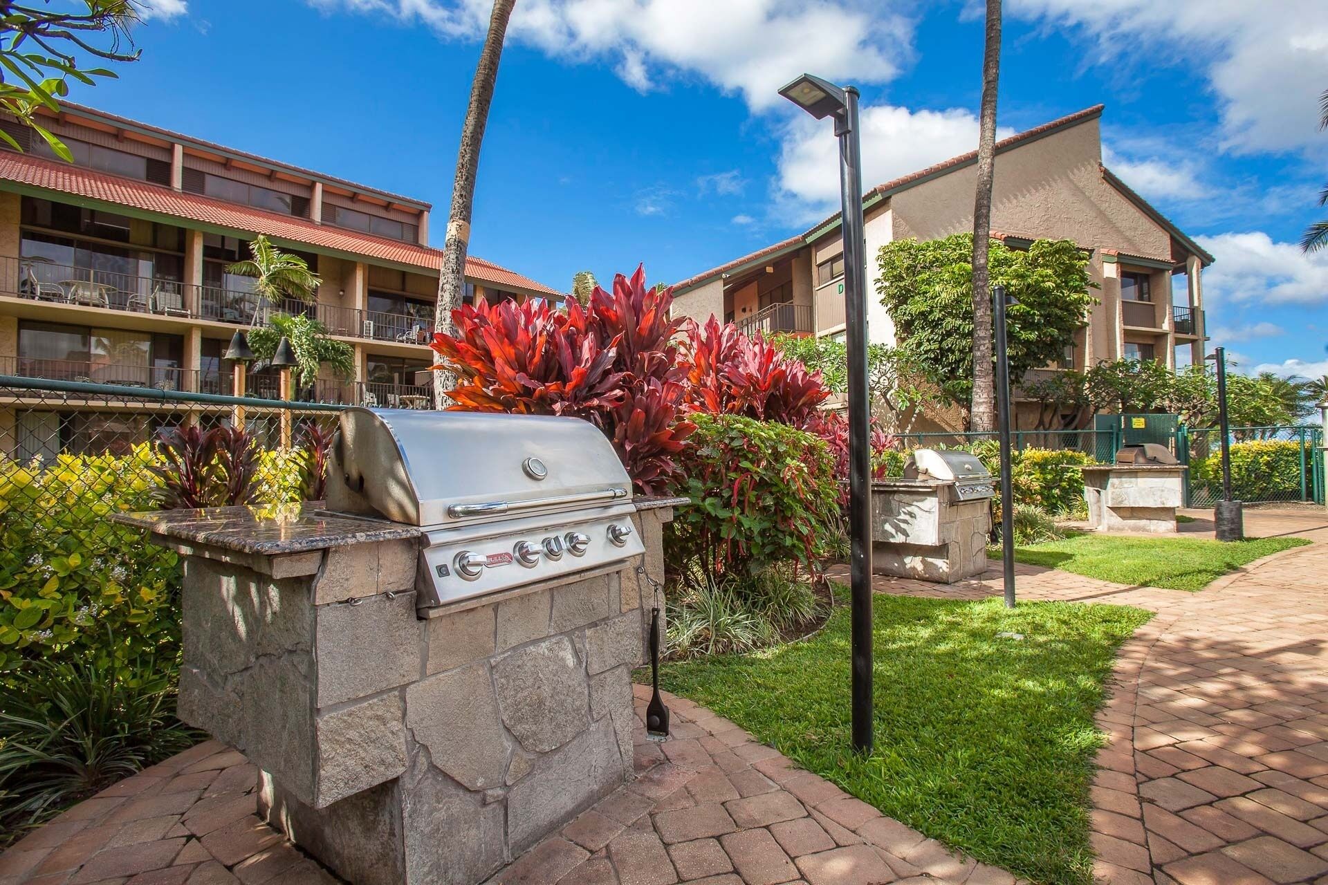 A stone barbecue grill with a gas burner, surrounded by lush greenery and a paved walkway.