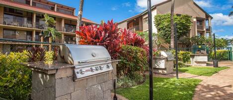 A stone barbecue grill with a gas burner, surrounded by lush greenery and a paved walkway.