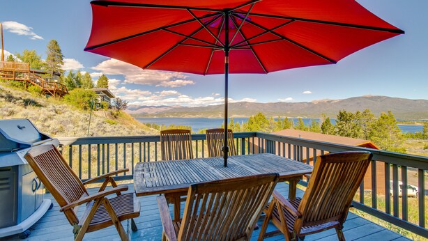 Une terrasse avec une table et des chaises, un parasol rouge et une vue sur les montagnes et l’eau.