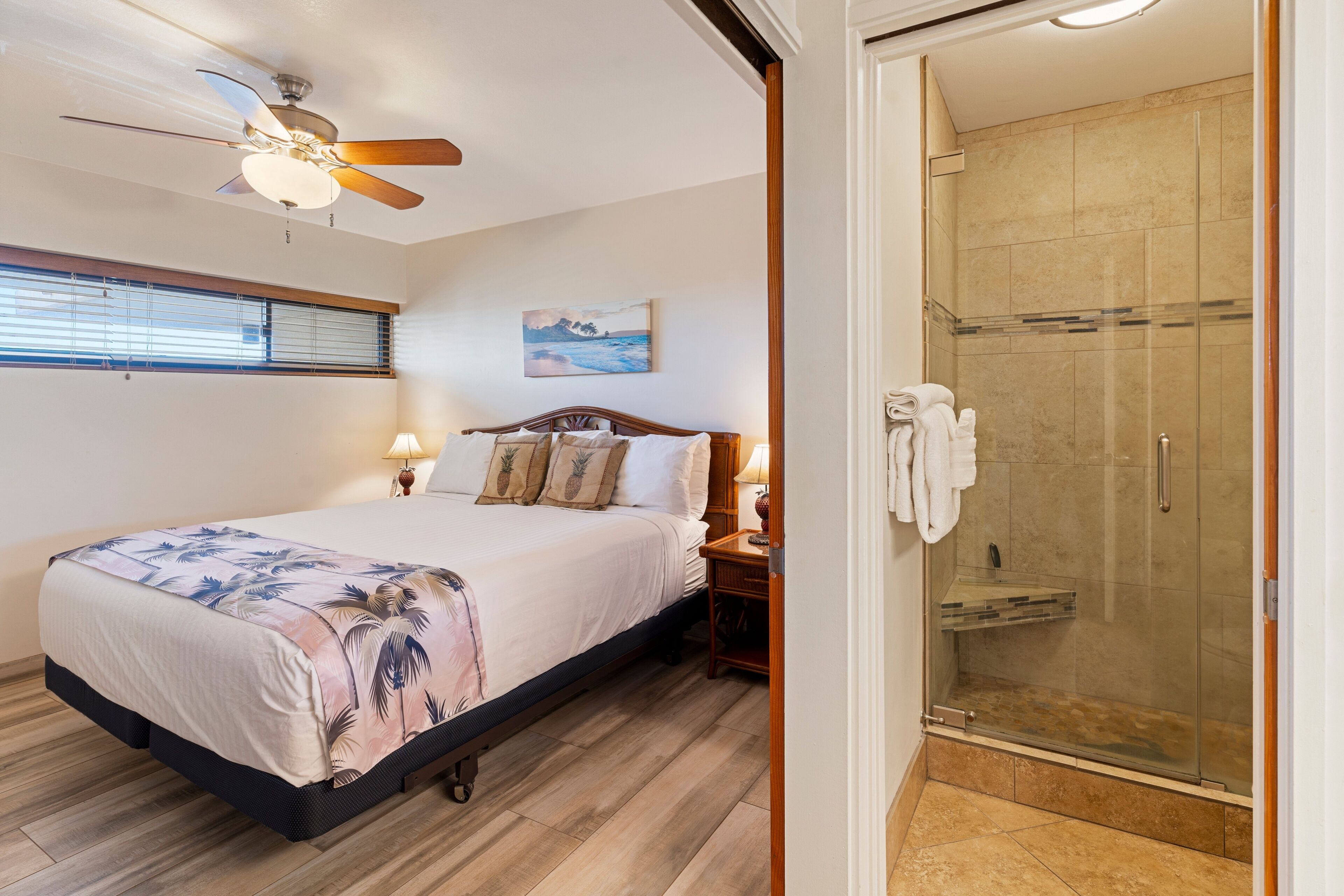 Hotelkamer met een bed, een plafondventilator, een schilderij, een nachtkastje, een lamp en een douche met glazen wanden.