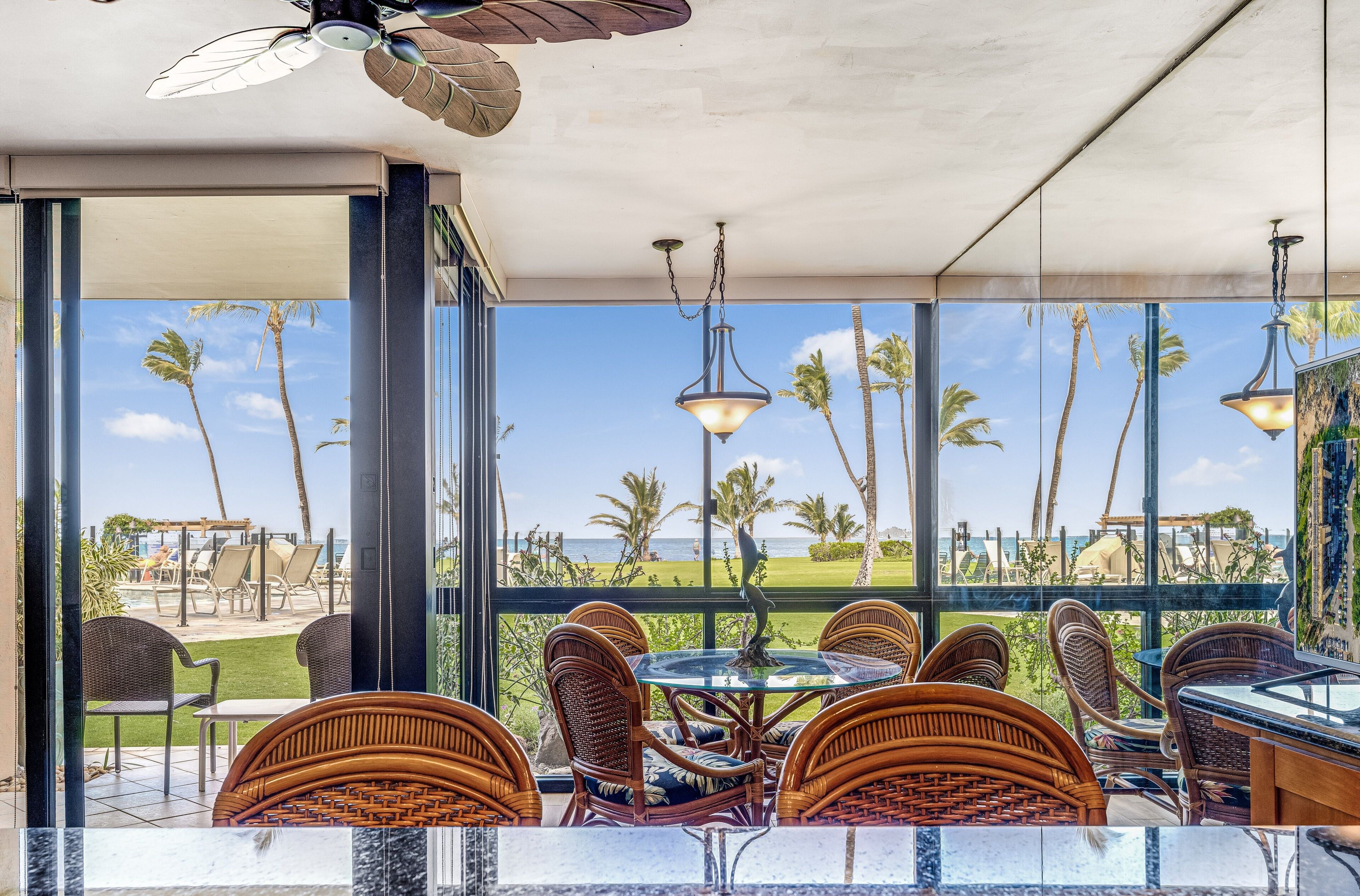 A dining area with wicker chairs and a glass table, overlooking a pool and palm trees.