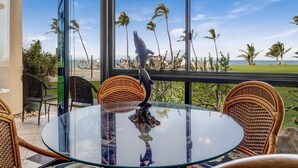 Une table ronde en verre, entourée de chaises en osier, avec vue sur une plage bordée de palmiers et l’océan.