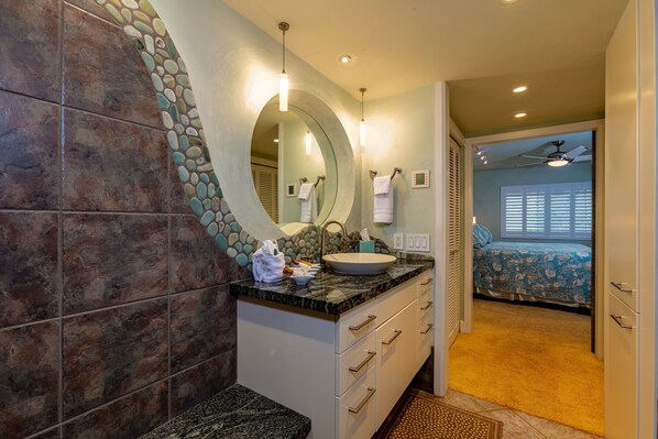 A bathroom with a stone wall, a sink, and a mirror. There is a towel rack and a shower area visible.