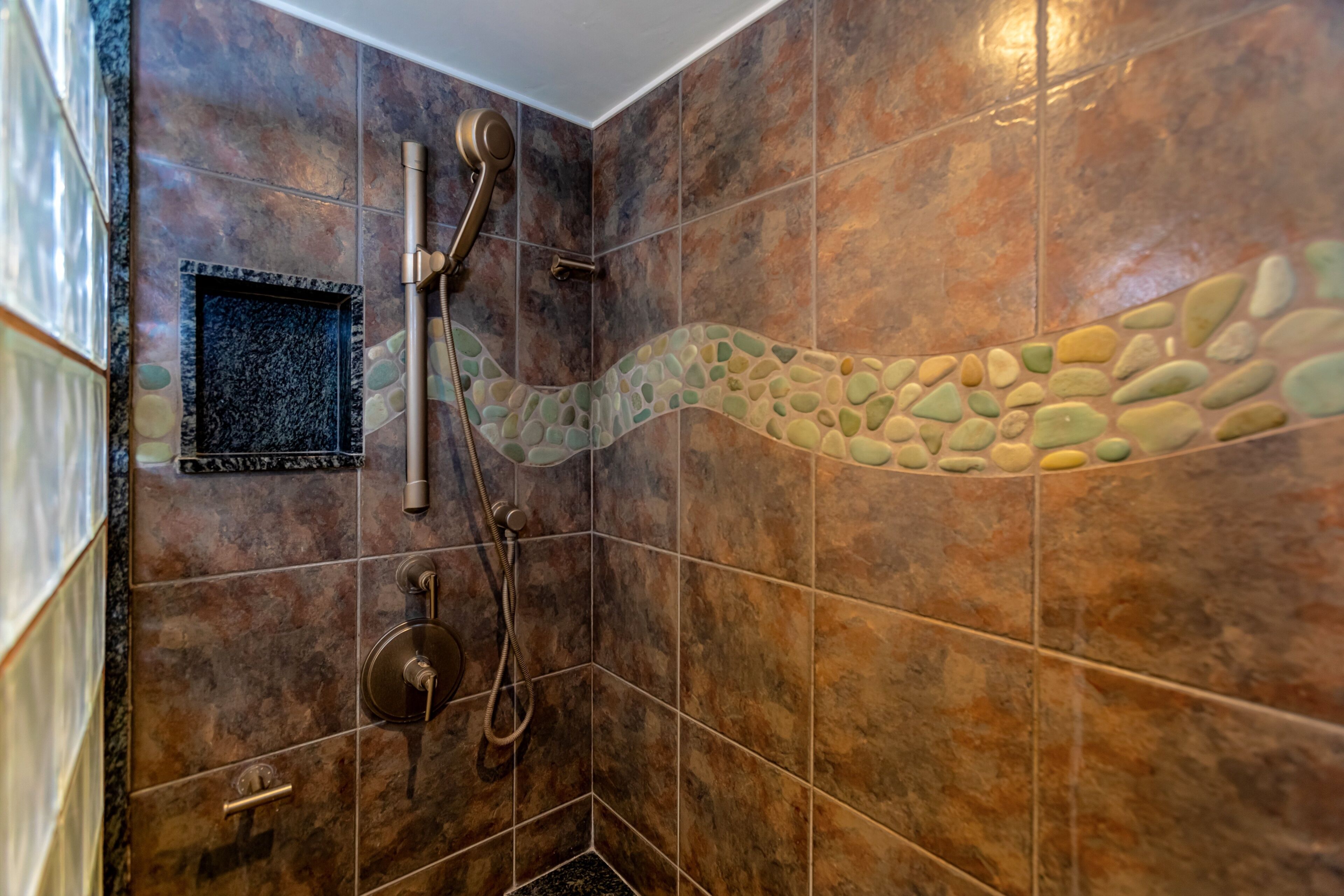 Un angolo doccia con mosaico in pietra sulla parete, un soffione a mano e un rubinetto fissato alla parete.