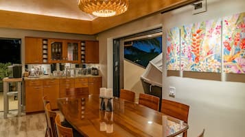 Uma cozinha moderna com armários de madeira, uma área de refeições com mesa de vidro e uma grande janela com vista para palmeiras.
