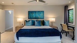 A hotel room with a bed, bedside lamps, a ceiling fan, a painting, a desk with a chair, and a window with curtains. - Makena Surf, #g-301-302 4 Bedroom Condo by RedAwning (Kihei)