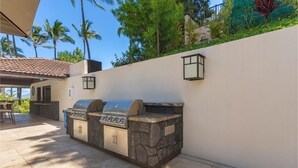 An outdoor kitchen area with a stone barbecue and a grill, surrounded by a white wall and palm trees. - Makena Surf, #g-301-302 4 Bedroom Condo by RedAwning (Kihei)
