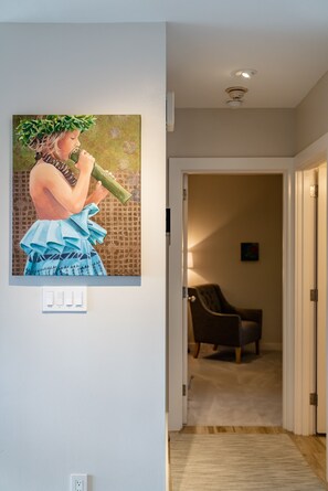 Un couloir orné d’un tableau représentant une personne tenant une bouteille, une chaise, et une porte donnant sur une autre pièce.
