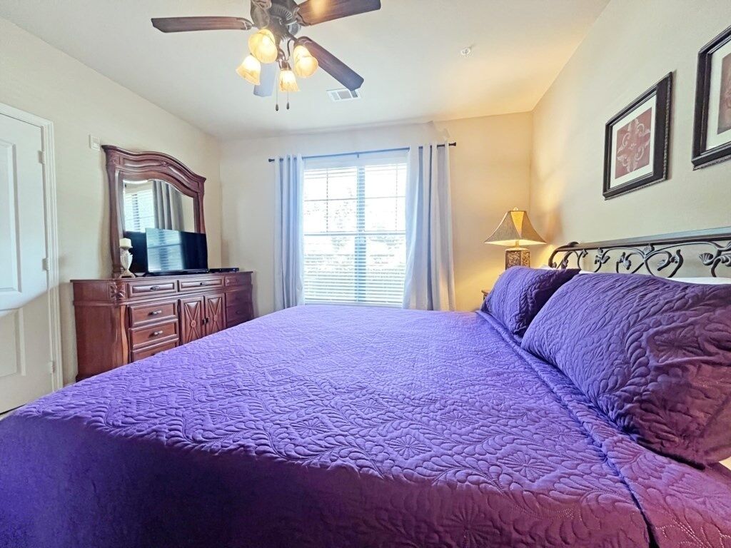 Une chambre à coucher avec un grand lit, un ventilateur de plafond, une commode et une fenêtre avec des rideaux.