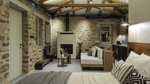 A rustic room with exposed wooden beams, stone walls, a fireplace, a sofa, a bed, and a small kitchen area.