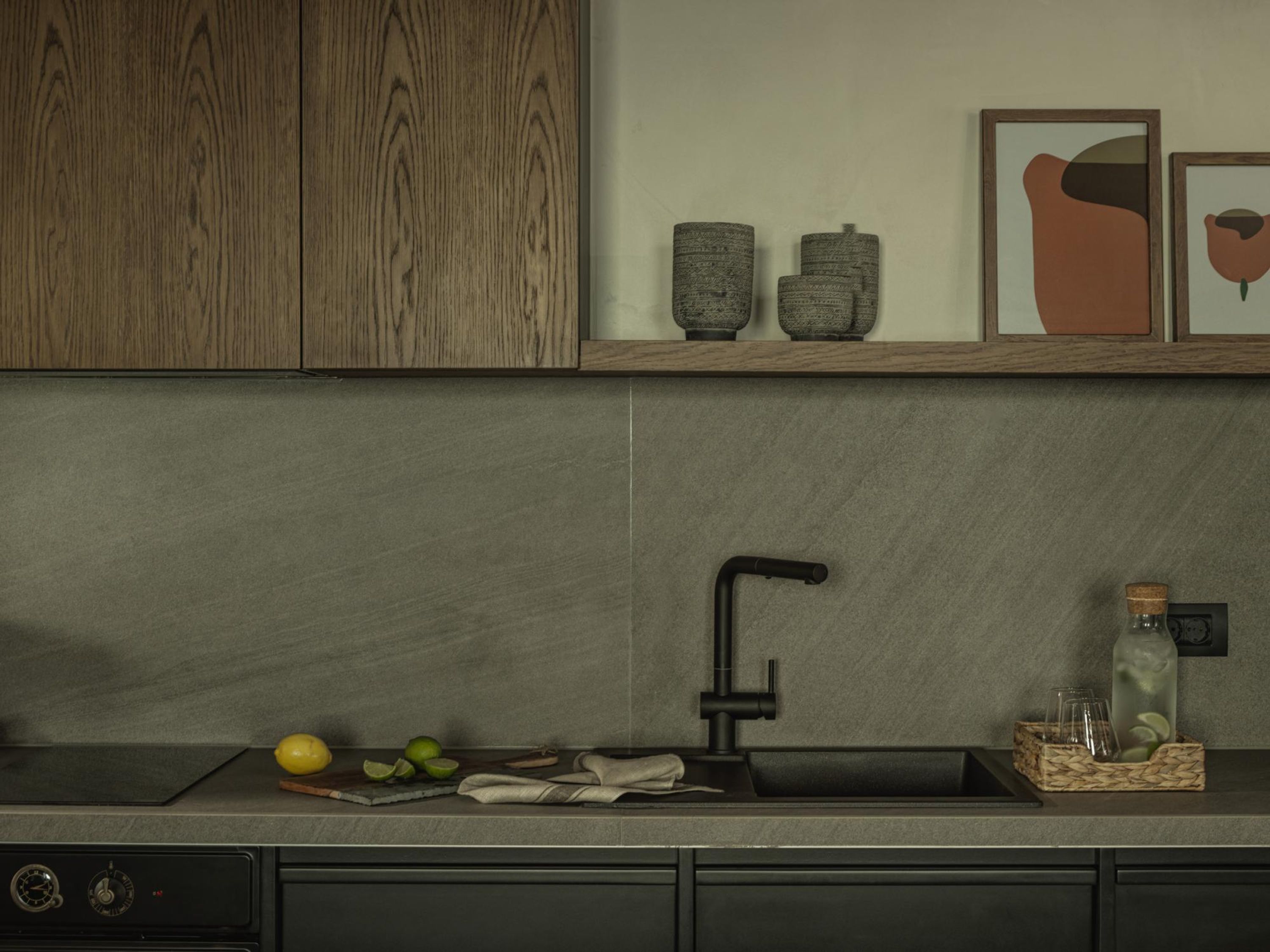 A modern kitchen with a grey countertop, wooden cabinets, a black faucet, and a small sink. There are decorative items on the shelf above the sink, including a framed picture and two ceramic vases.