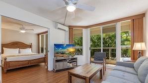 A hotel room with a bed, a TV, a sofa, a coffee table, and large windows with a view of greenery. - Maui Banyan by Coldwell Banker Island Vacations (Kihei)