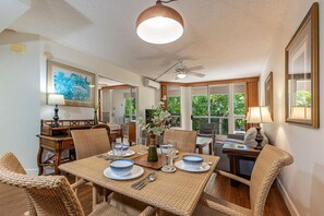 Un espace repas aménagé avec des meubles en osier, un ventilateur de plafond et de grandes fenêtres donnant sur un paysage verdoyant.