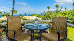 A balcony with wicker chairs, a round table, and a view of the ocean and palm trees. - Maui Banyan by Coldwell Banker Island Vacations (Kihei)