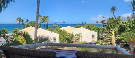 A balcony with wicker chairs overlooking a beach with palm trees and a clear blue sky.