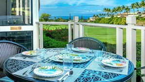 Una mesa de comedor dispuesta con platos, vasos y cubiertos en un balcón con vista a un campo de golf y al océano.