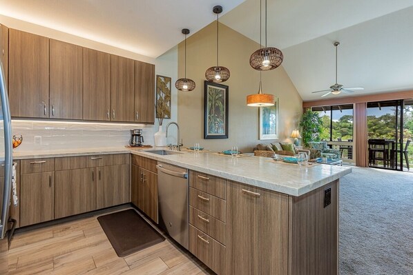 A modern kitchen with wooden cabinets, a central island, pendant lighting, and a view of the outdoors. - Grand Champions Two Bedrooms - Garden View by Coldwell Banker Island Vacations (Kihei)