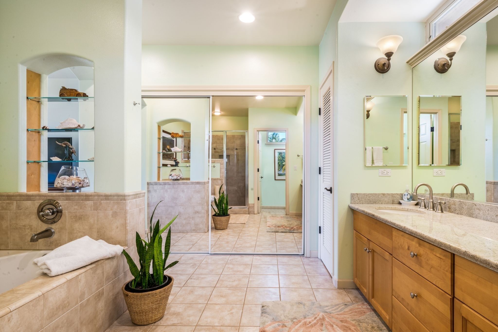 A bathroom with a bathtub, a sink with a mirror, and a shower area. There are shelves with decorative items and a potted plant.
