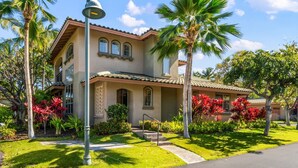 A two-story house with a tiled roof, a front porch, and a well-maintained garden with palm trees and red flowering plants. - Big Island Fairways At Mauna Lani 1701 2 Bedroom Condo (Kamuela)