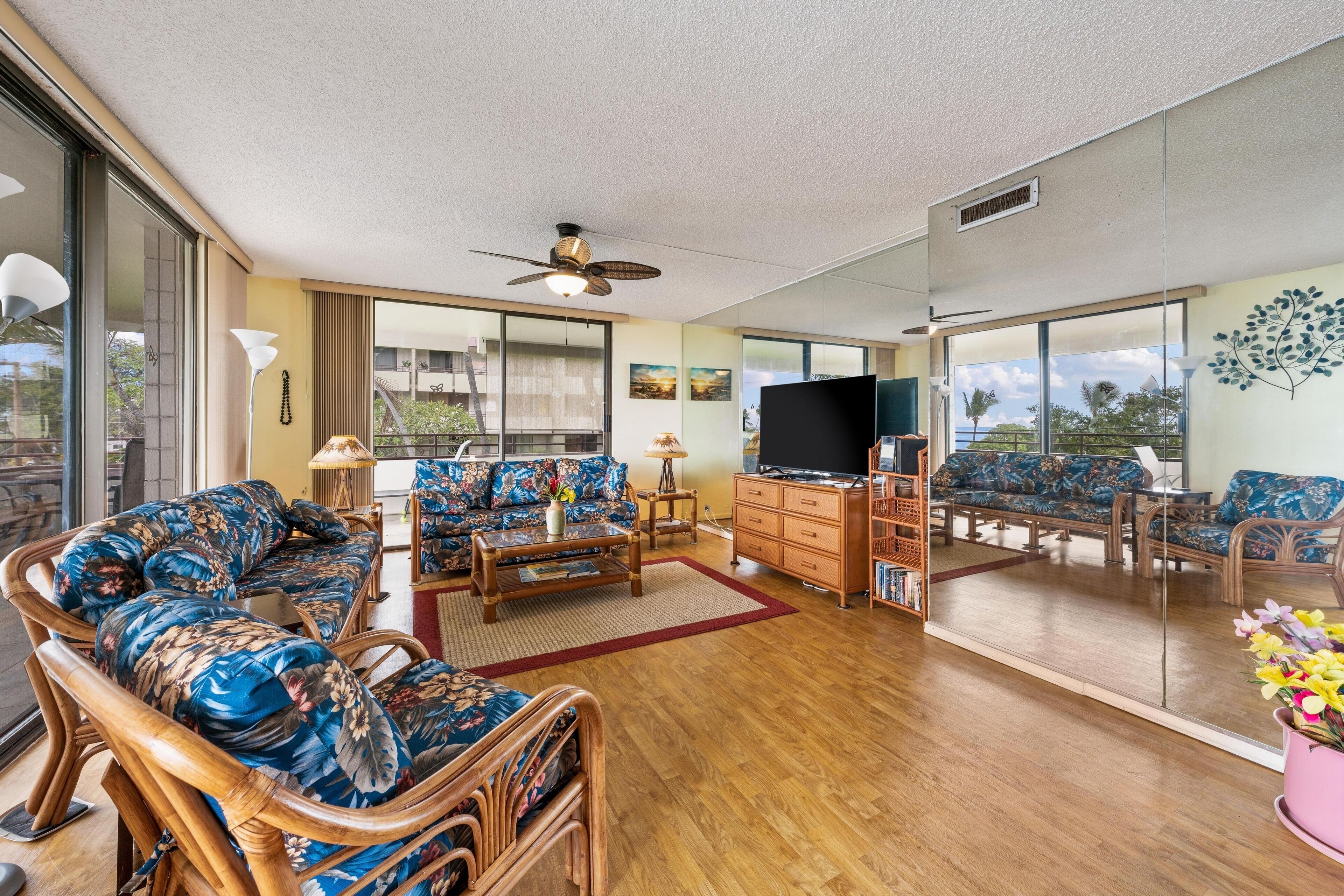 A spacious living room with a large flat-screen TV, wicker furniture, and a view of the outdoors through large windows.