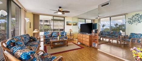 A spacious living room with a large flat-screen TV, wicker furniture, and a view of the outdoors through large windows.