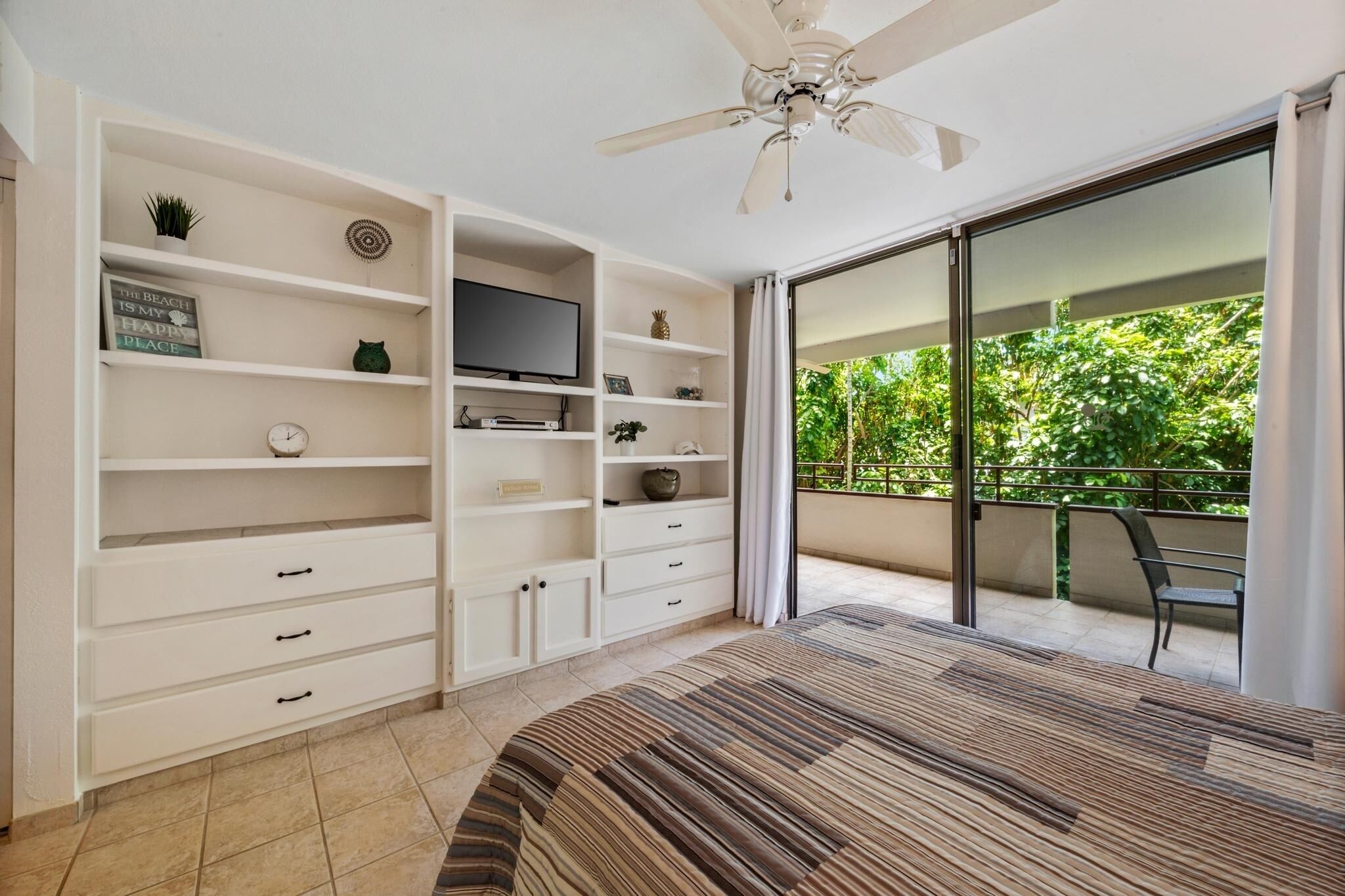 Une chambre avec un lit, une télévision, un ventilateur de plafond, un balcon avec une chaise et une vue sur la verdure.