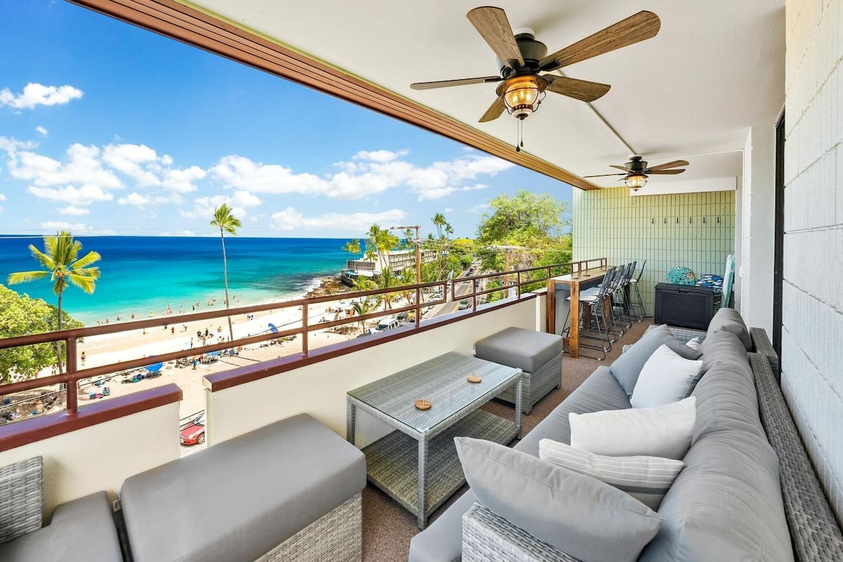 Ein Balkon mit Blick auf einen Strand, einer Couch, einem Tisch und Deckenventilatoren.