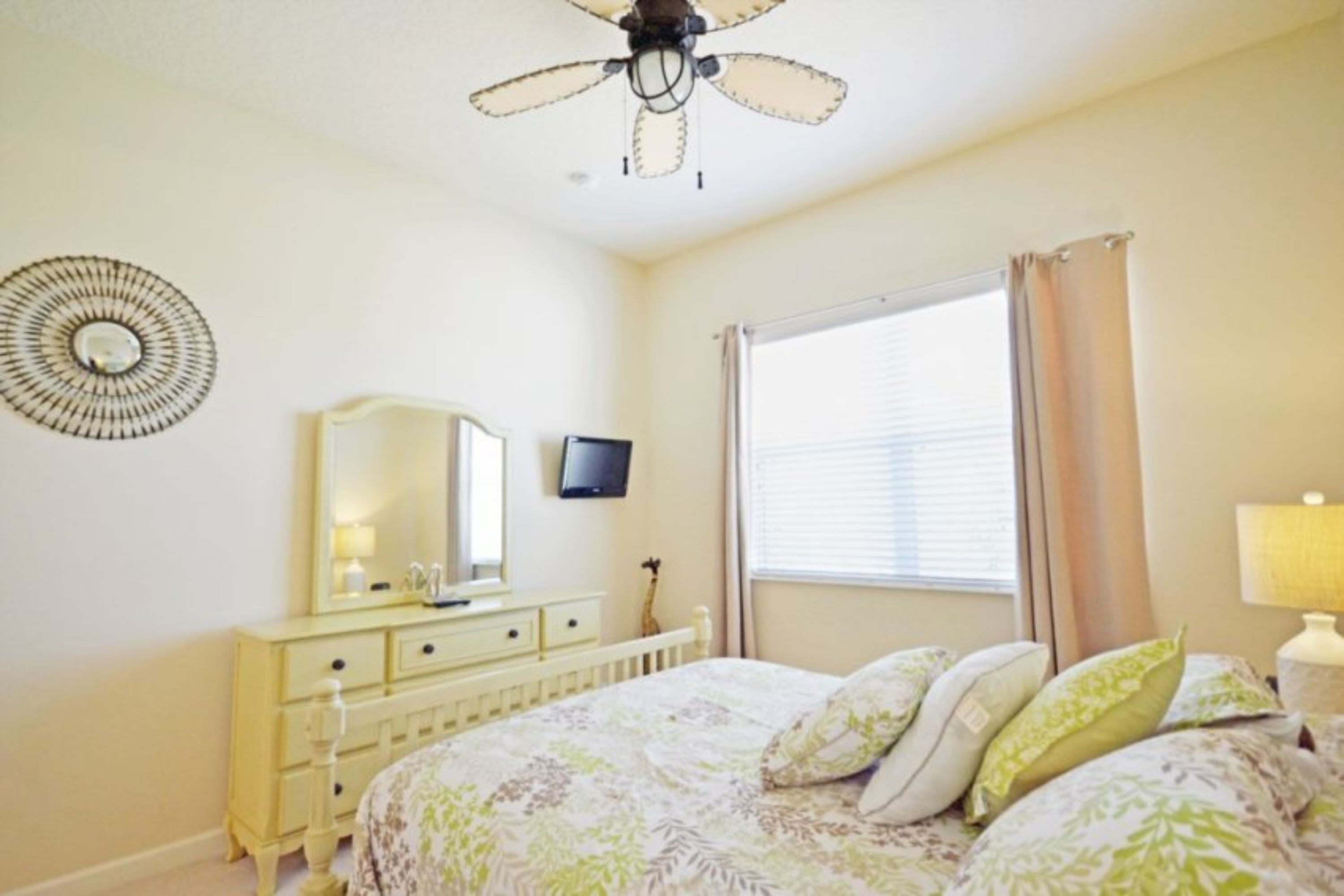 A bedroom with a bed, a dresser, a mirror, a ceiling fan, a window with curtains, and a wall decoration.